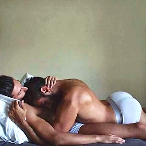 Two men nuzzle in bed.
